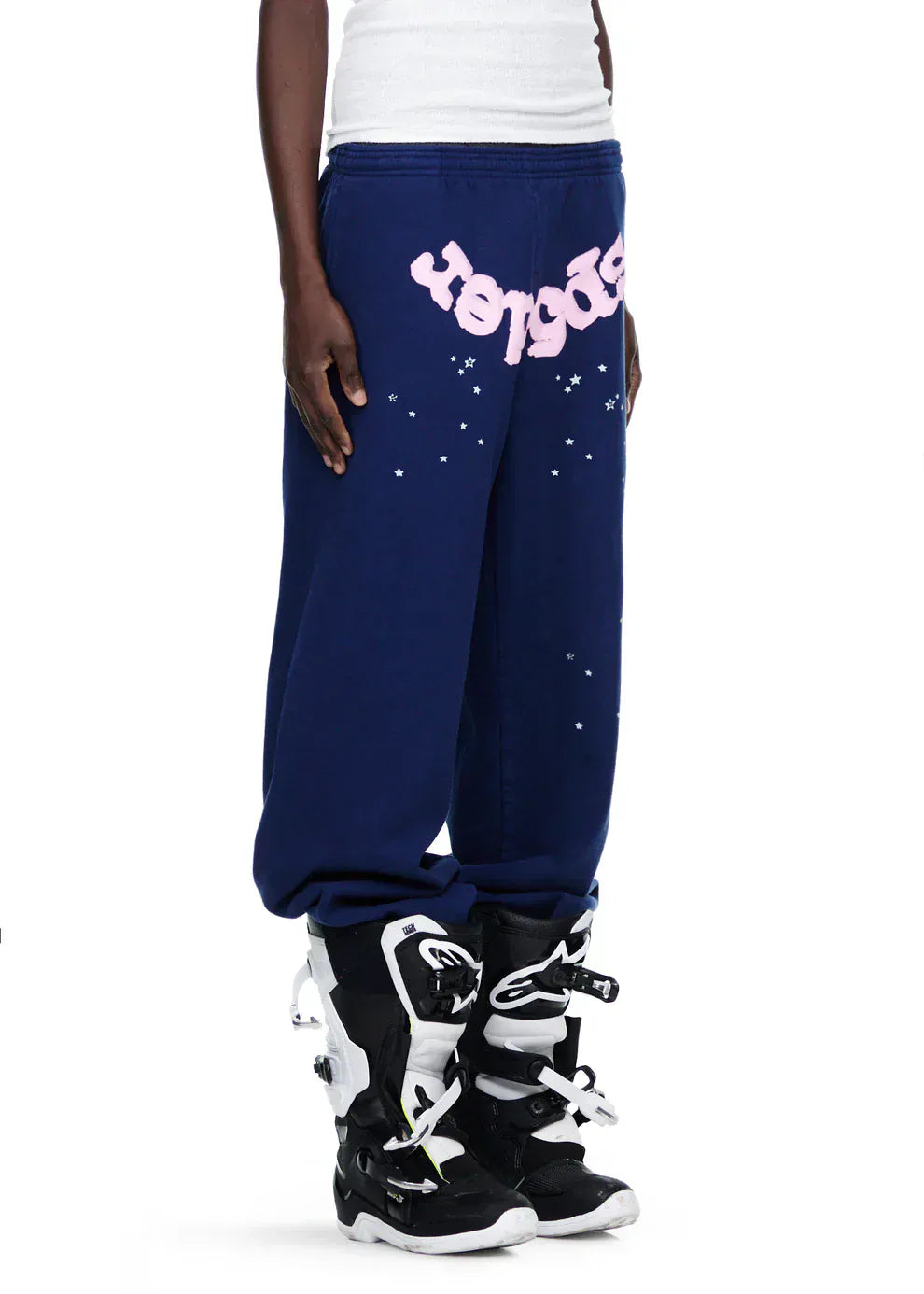 A person in Sp5der Sweatpants—unisex navy blue cotton joggers with a pink front logo and star print—paired with black and white motocross boots and a white shirt for a bold, sporty streetwear look.
