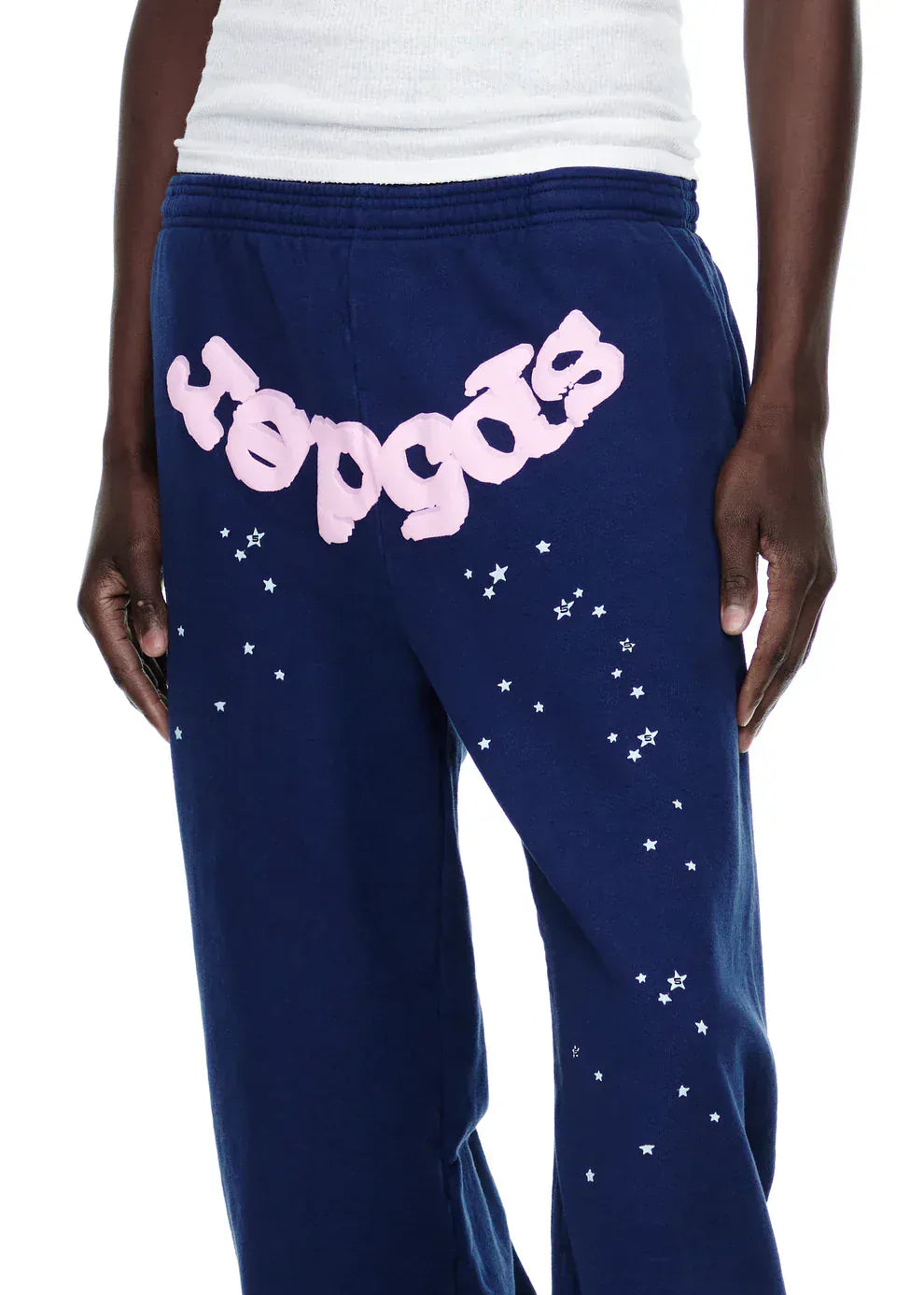 A person wearing Sp5der Sweatpants—unisex navy blue cotton sweatpants with a pink front logo and star print—stands with one hand by their side.