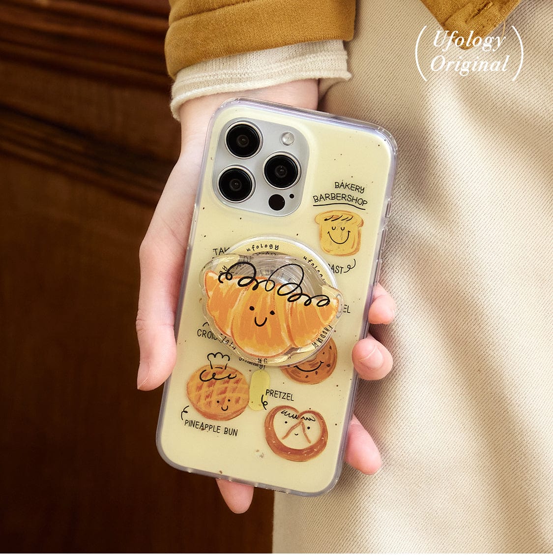 A person is holding a Bakery Friends MagSafe iPhone 16 Pro Max Case with an all-inclusive transparent protective cover featuring cute, illustrated baked goods like pretzels and buns, each with smiling faces. The case includes the text "Ufology Original" at the upper center and accommodates the phone’s three camera lenses.