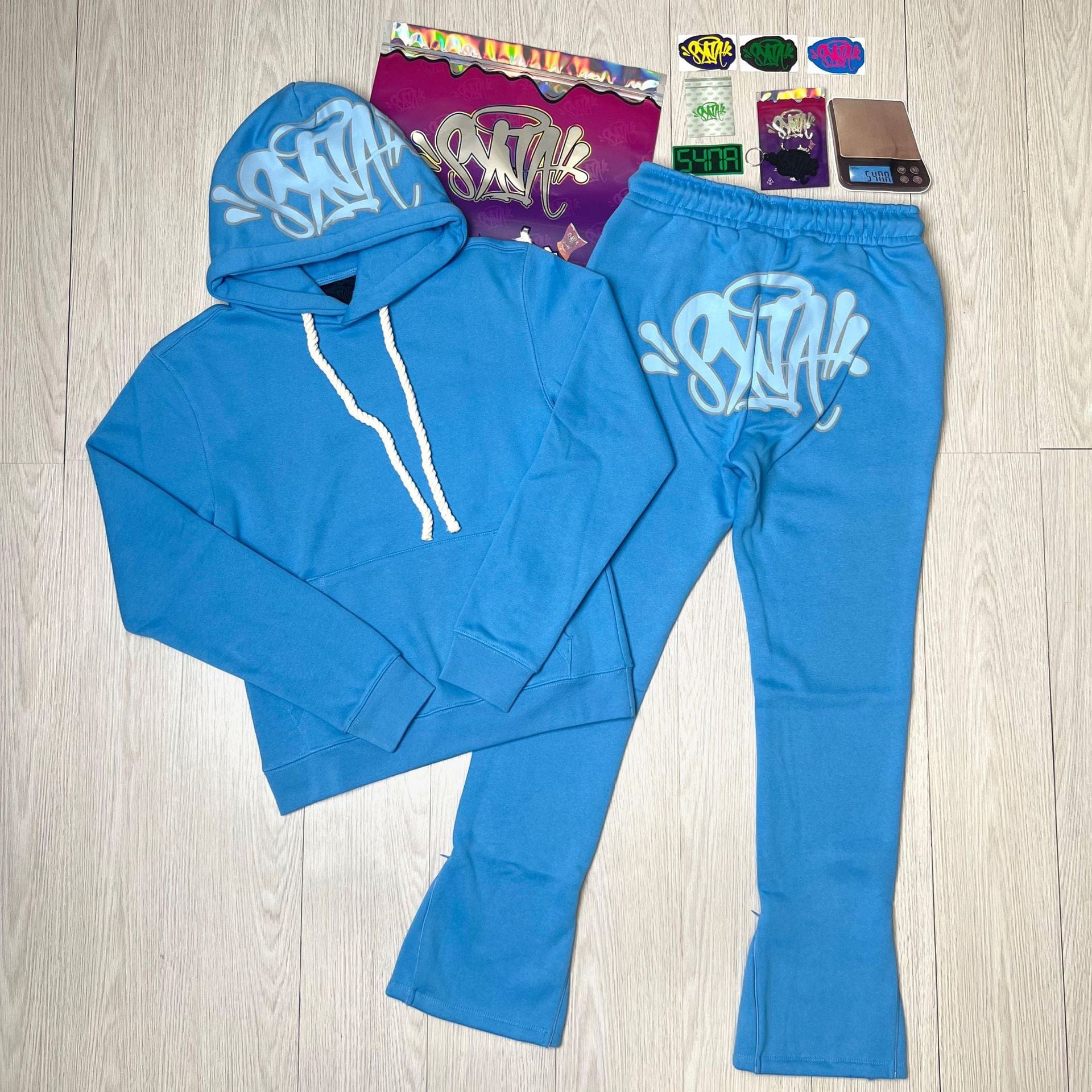 The Syna World Tracksuit Set – Graphic Hoodie & Flared Pants with Accessories features bold white graffiti graphics and comes with five stickers, a colorful bag, and a small digital scale, all shown on a light wood floor.