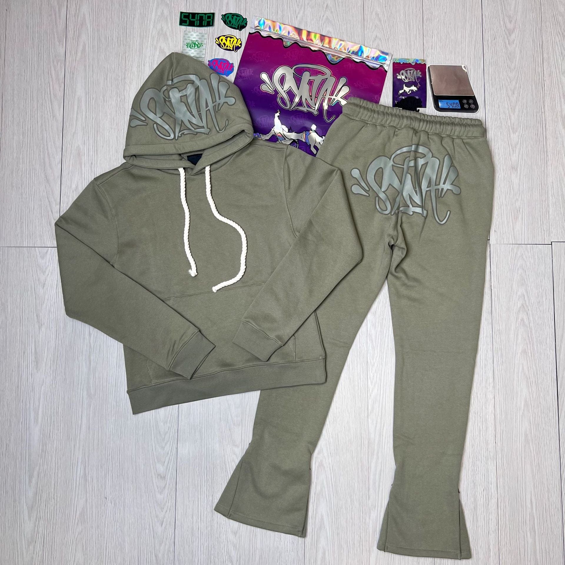 The Syna World Tracksuit Set – Graphic Hoodie & Flared Pants with Accessories in olive green, featuring gray graffiti text, is displayed on a light floor with stickers, packaging, and a small scale in the background.