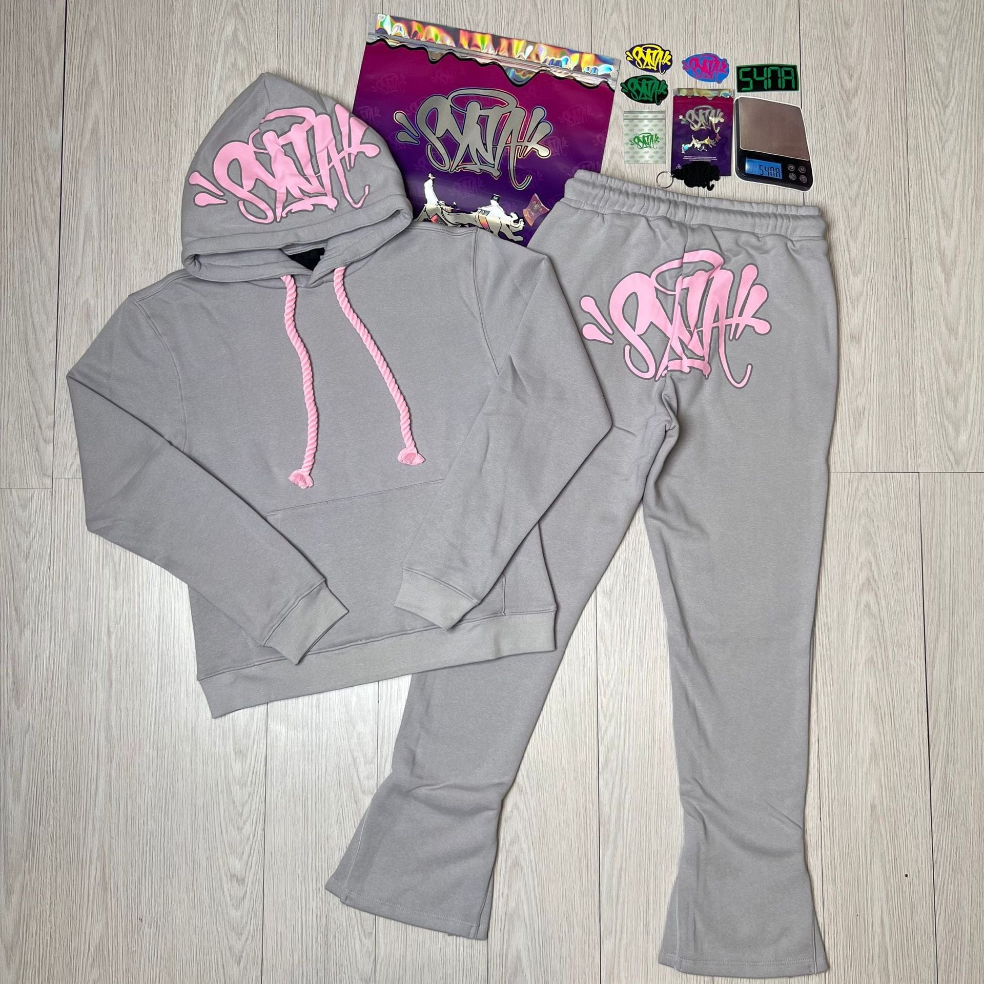 The Syna World Tracksuit Set features a gray graphic hoodie and flared pants with pink graffiti-style text, displayed on a light wood floor beside colorful stickers and cards included as accessories.