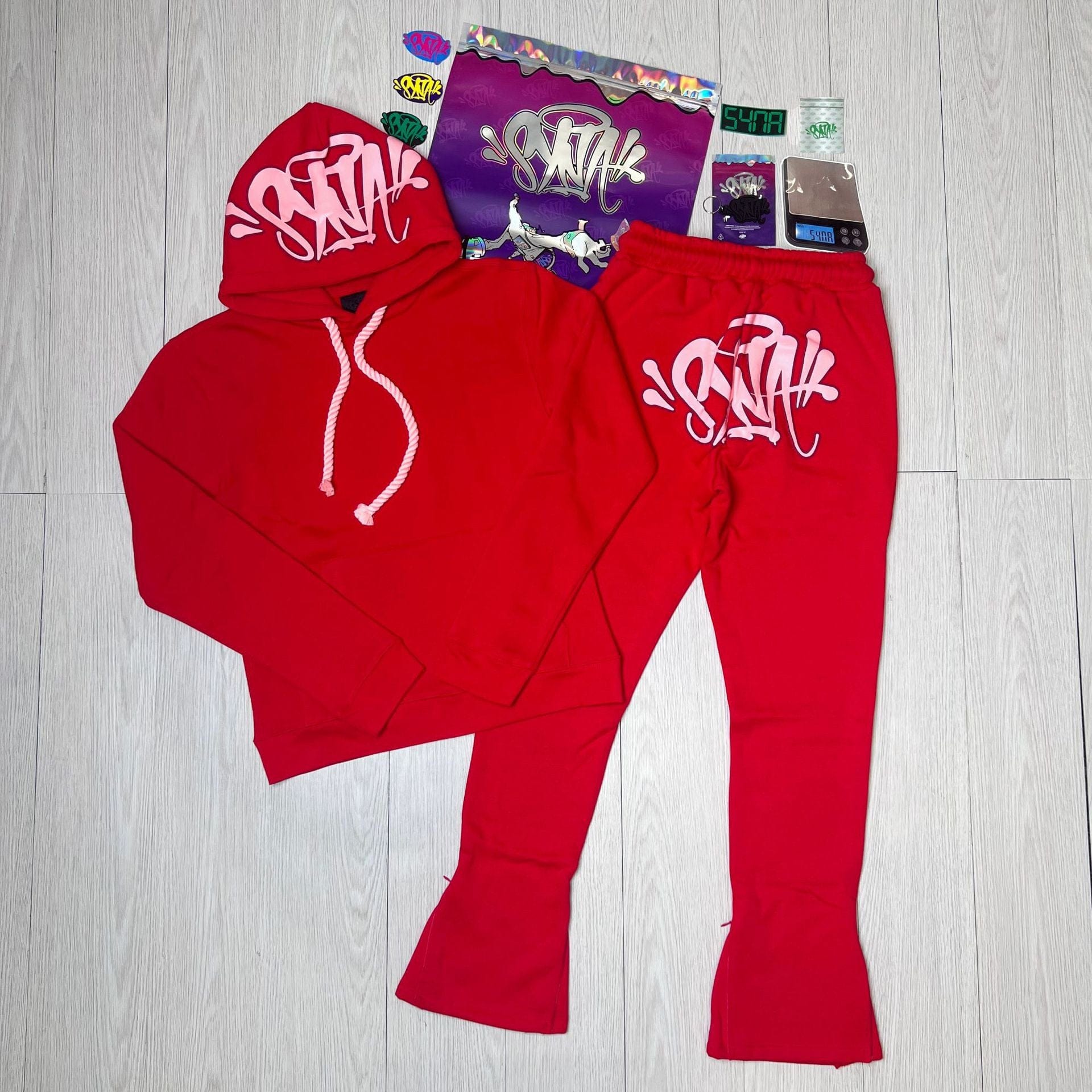 The Syna World Tracksuit Set features a red graphic hoodie and flared pants with white graffiti text, plus accessories, all displayed on a light wood floor with assorted stickers—showcasing iconic Syna World clothing.