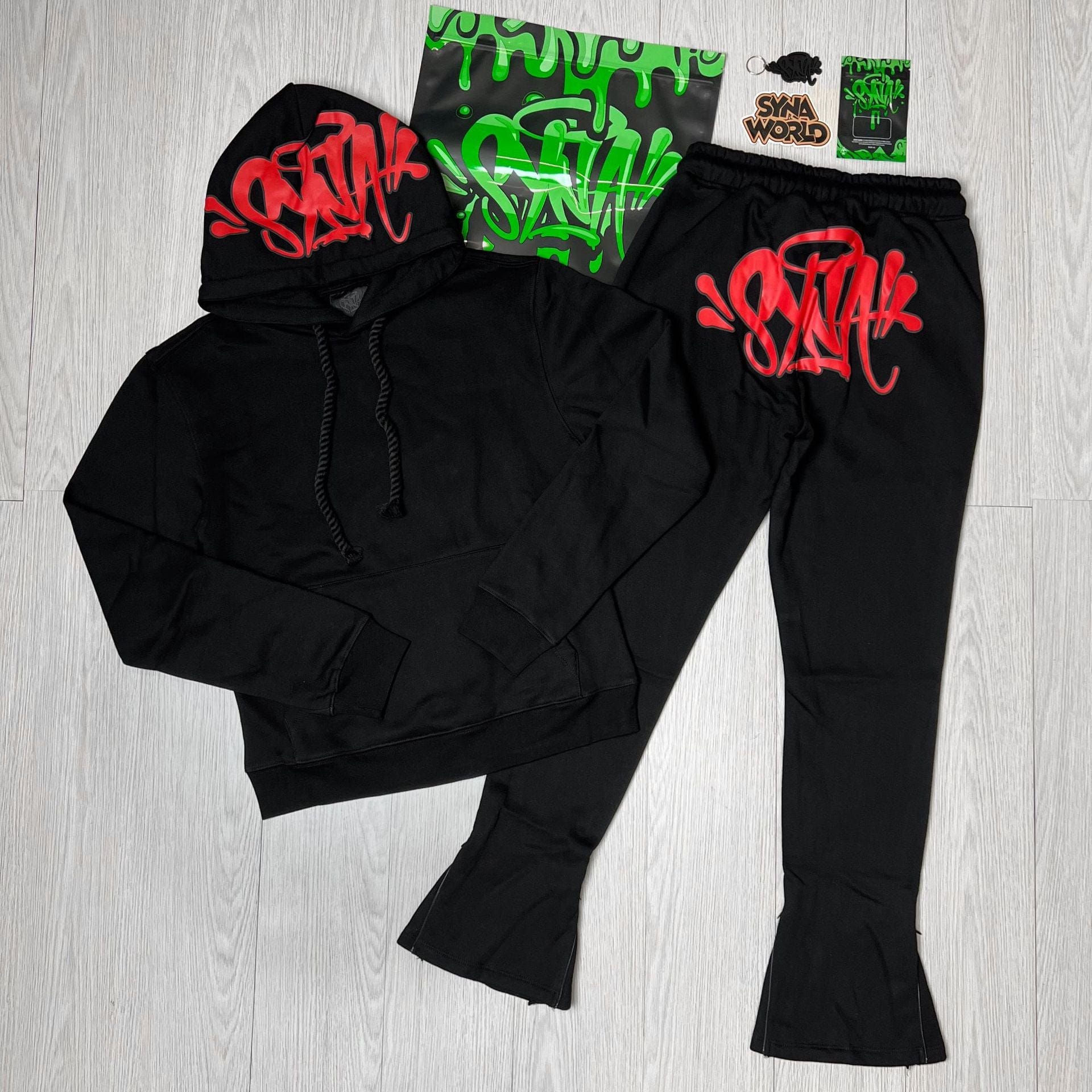 The Syna World Graphic Hoodie and Flared Pants Tracksuit Set with Accessories comes in 4 colors, featuring graffiti-style graphics and matching stickers and a pin, displayed on a light wood floor.