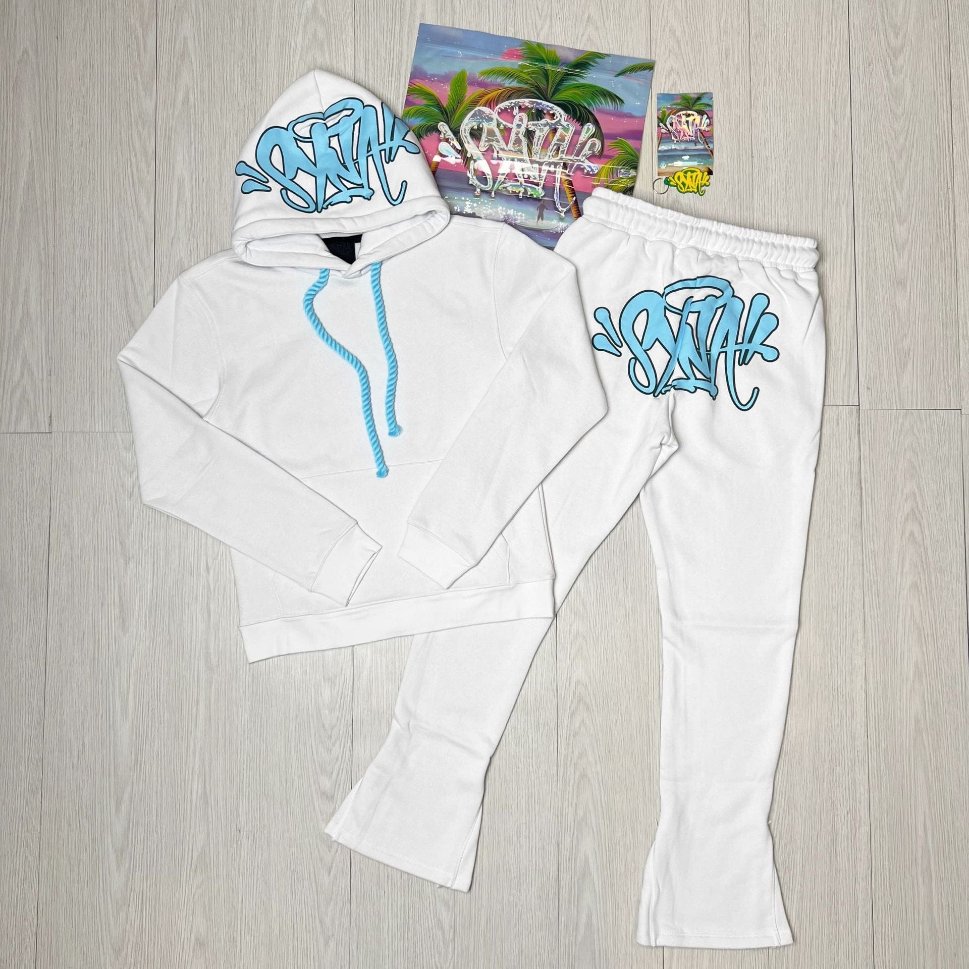 The Syna World Tracksuit Set – Graphic Hoodie & Flared Pants with Accessories, featuring blue graffiti-style text, is displayed on a light wood floor alongside a vibrant album cover for a bold fashion statement.
