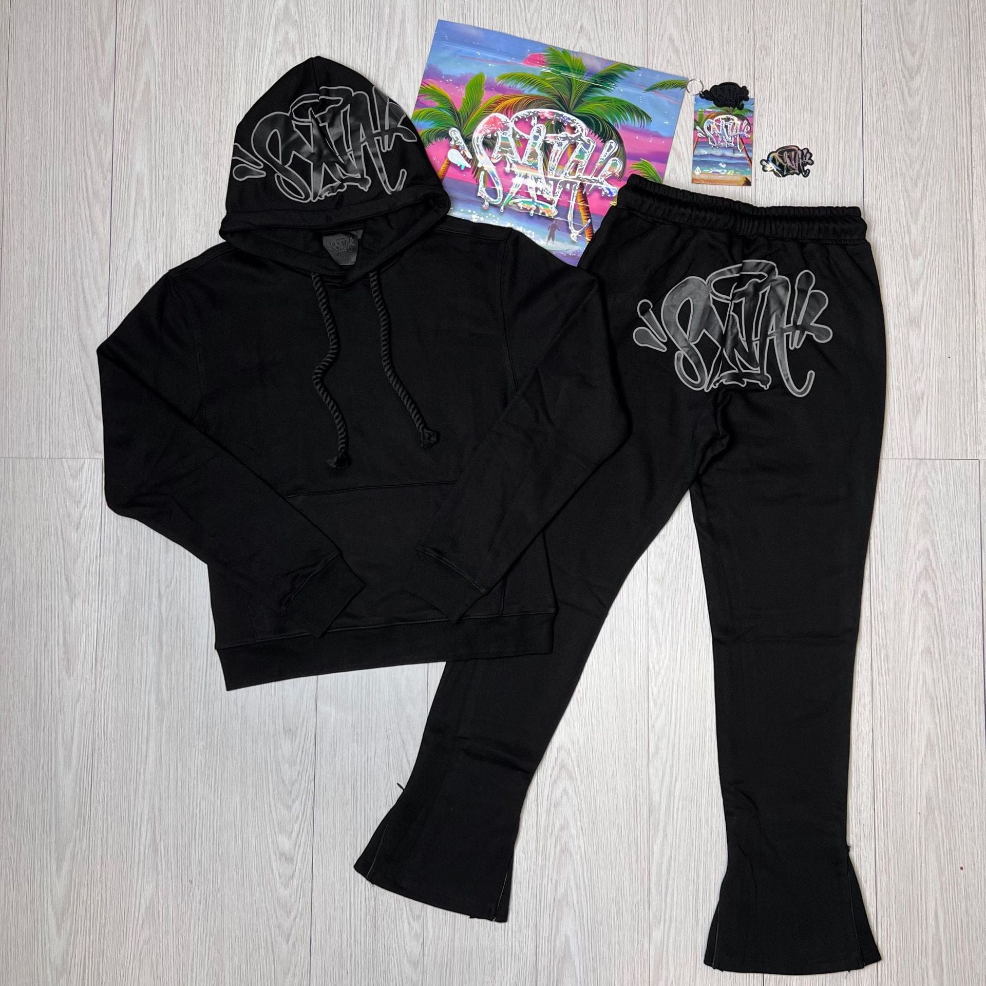 Syna World Tracksuit Set—Graphic Hoodie & Flared Pants with Accessories—featuring white graffiti text, displayed on a light wood floor next to colorful items with a tropical print.
