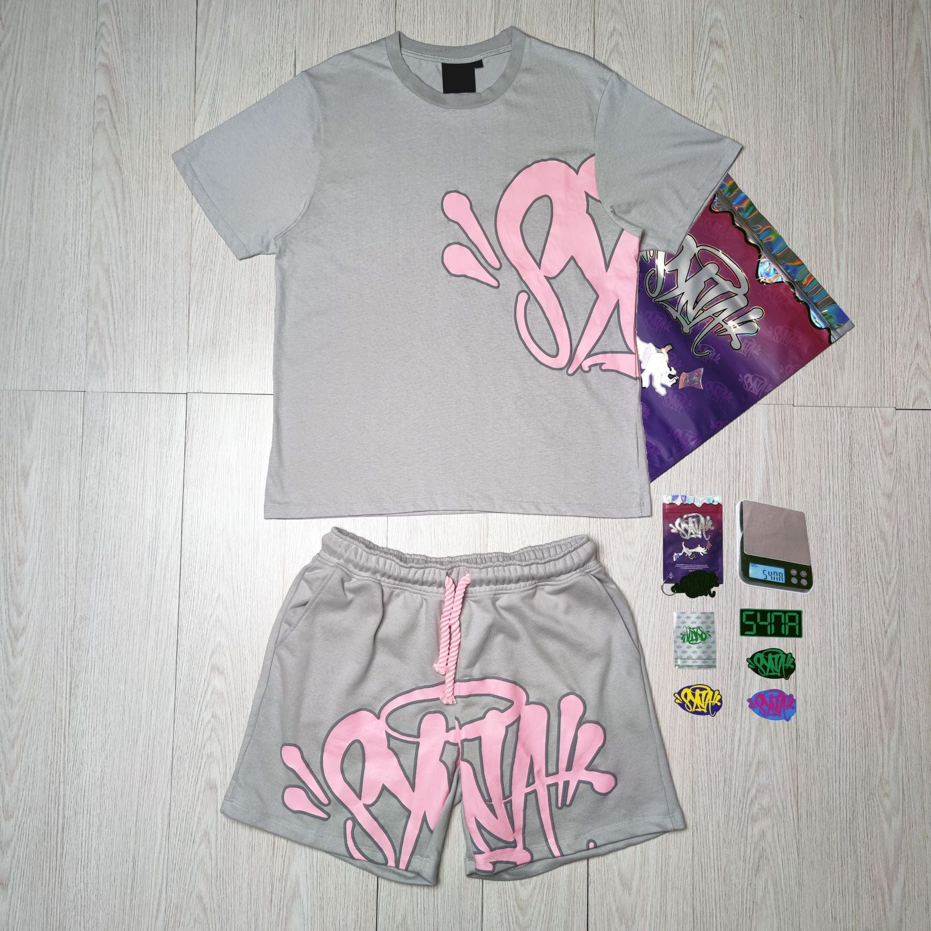 The Syna World T-Shirt and Shorts Set with Accessories – 8 Colors is displayed on a light wood floor alongside colorful stickers, a gray wallet, and a decorative card—an ideal pick for standout streetwear style.