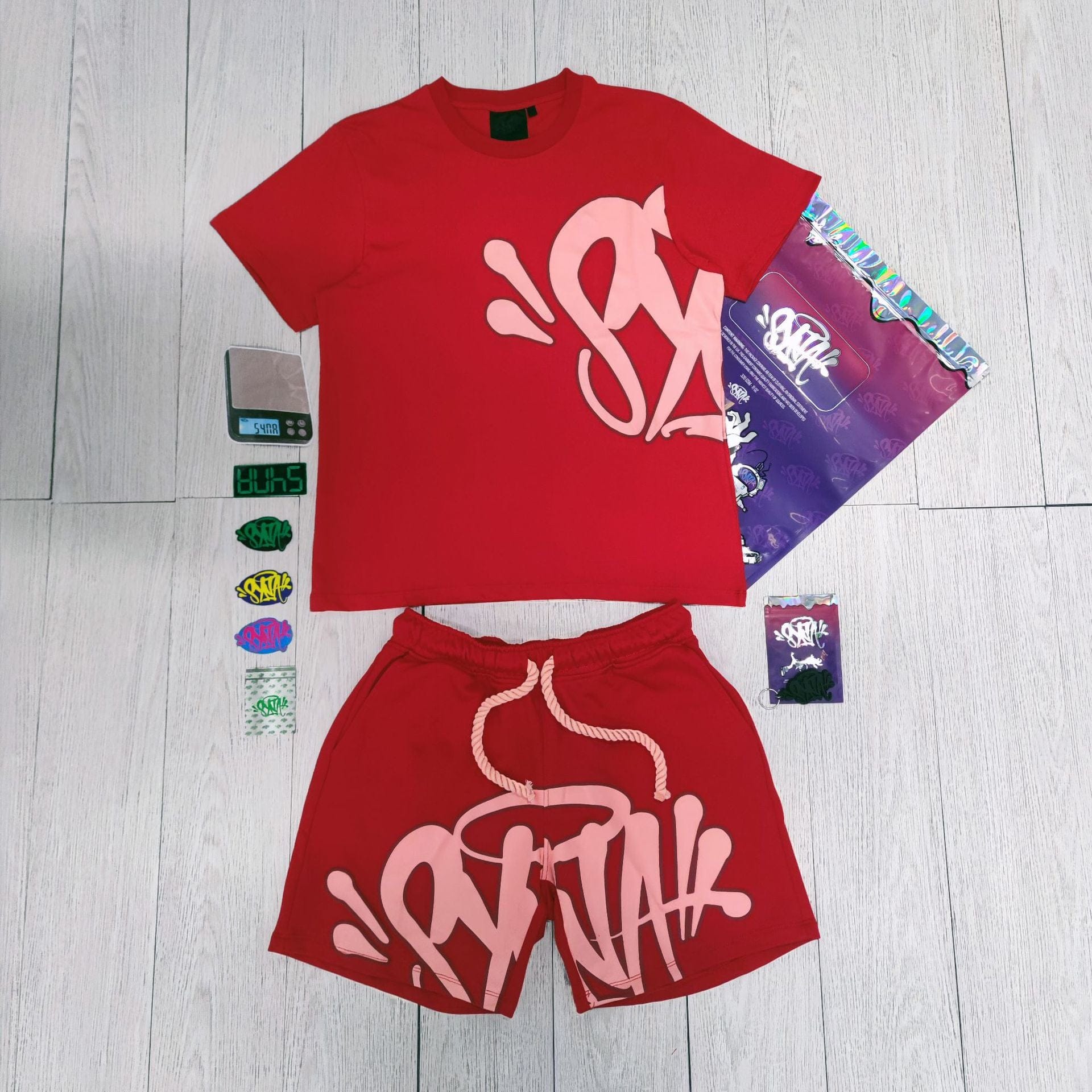 The Syna World T-Shirt and Shorts Set with Accessories in red, featuring bold white graffiti-style lettering, stands out as a unique streetwear outfit—shown with stickers, packaging, and a digital scale on a light wood floor.