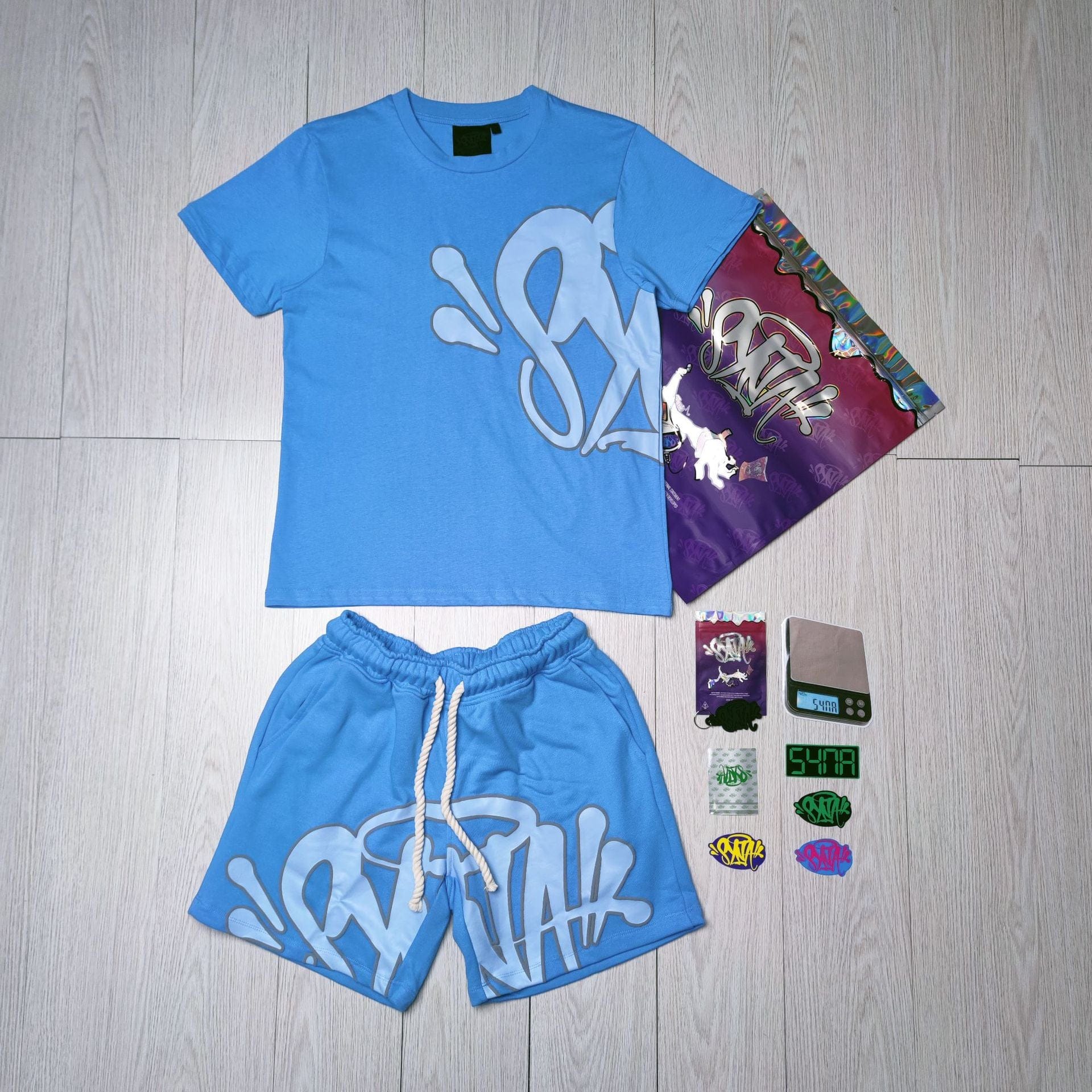 The Syna World T-Shirt and Shorts Set with Accessories – 8 Colors is displayed in blue with graffiti-style text as a trendy streetwear outfit, paired with a branded bag, tags, and stickers on a light wood floor.