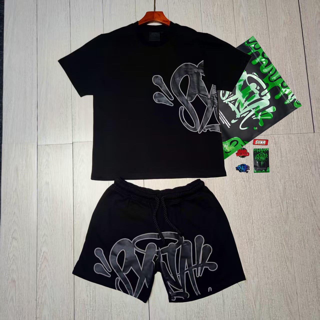 The Syna World T-Shirt and Shorts Set with Accessories – 8 Colors features bold silver graffiti-style lettering and is displayed on a light wood floor with matching stickers and tags beside the streetwear outfit.
