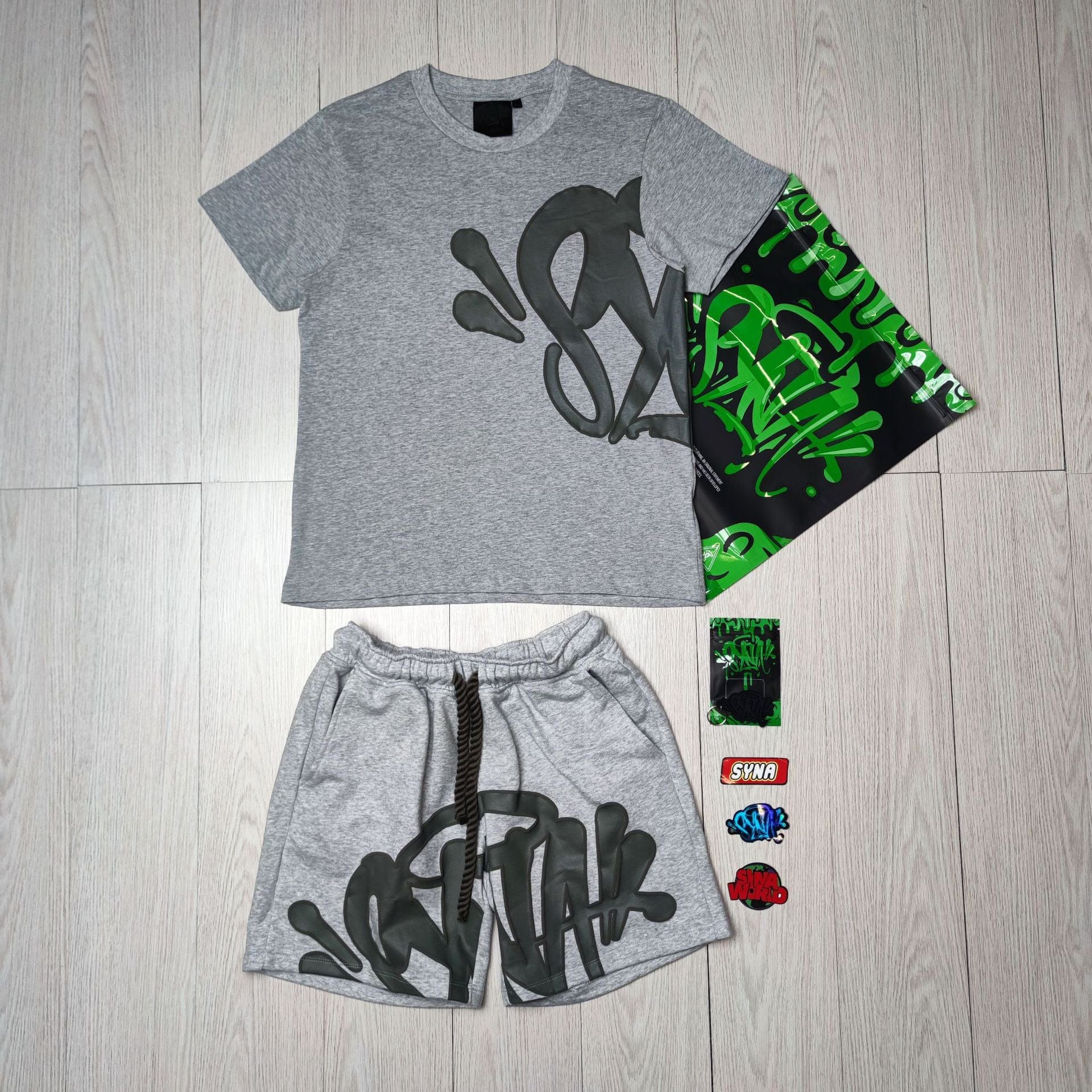 The Syna World T-Shirt and Shorts Set with Accessories – 8 Colors features gray pieces with black and green graffiti-style lettering, a matching green and black bag, plus four assorted streetwear pins or patches.