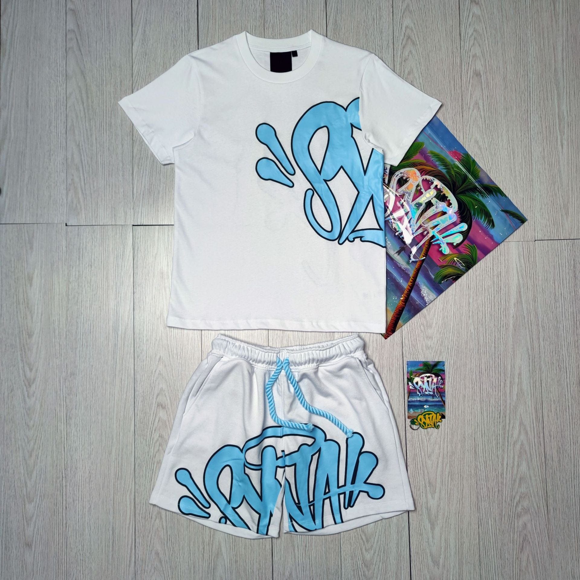Syna World T-Shirt and Shorts Set with Accessories – 8 Colors, shown in white with blue graffiti design on a light wood floor beside a colorful poster and sticker packs—ideal for upgrading your streetwear style.