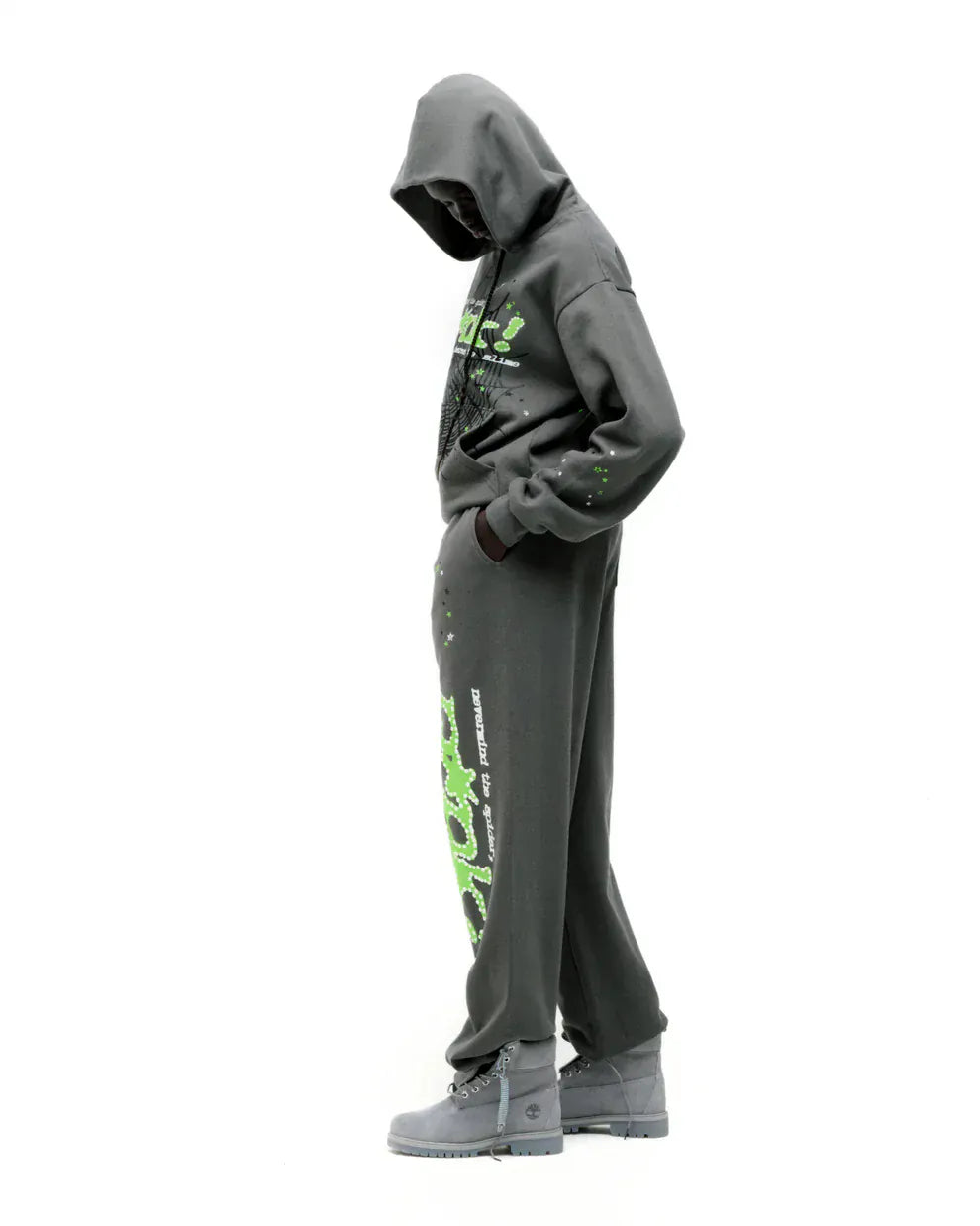 Wearing the Sp5der “P!nk! Hares the Slime” Olive Green 440GSM Heavyweight Spider-Web Pullover hoodie, this person stands sideways against a white background—hands in pockets and head down—for a sleek streetwear vibe.