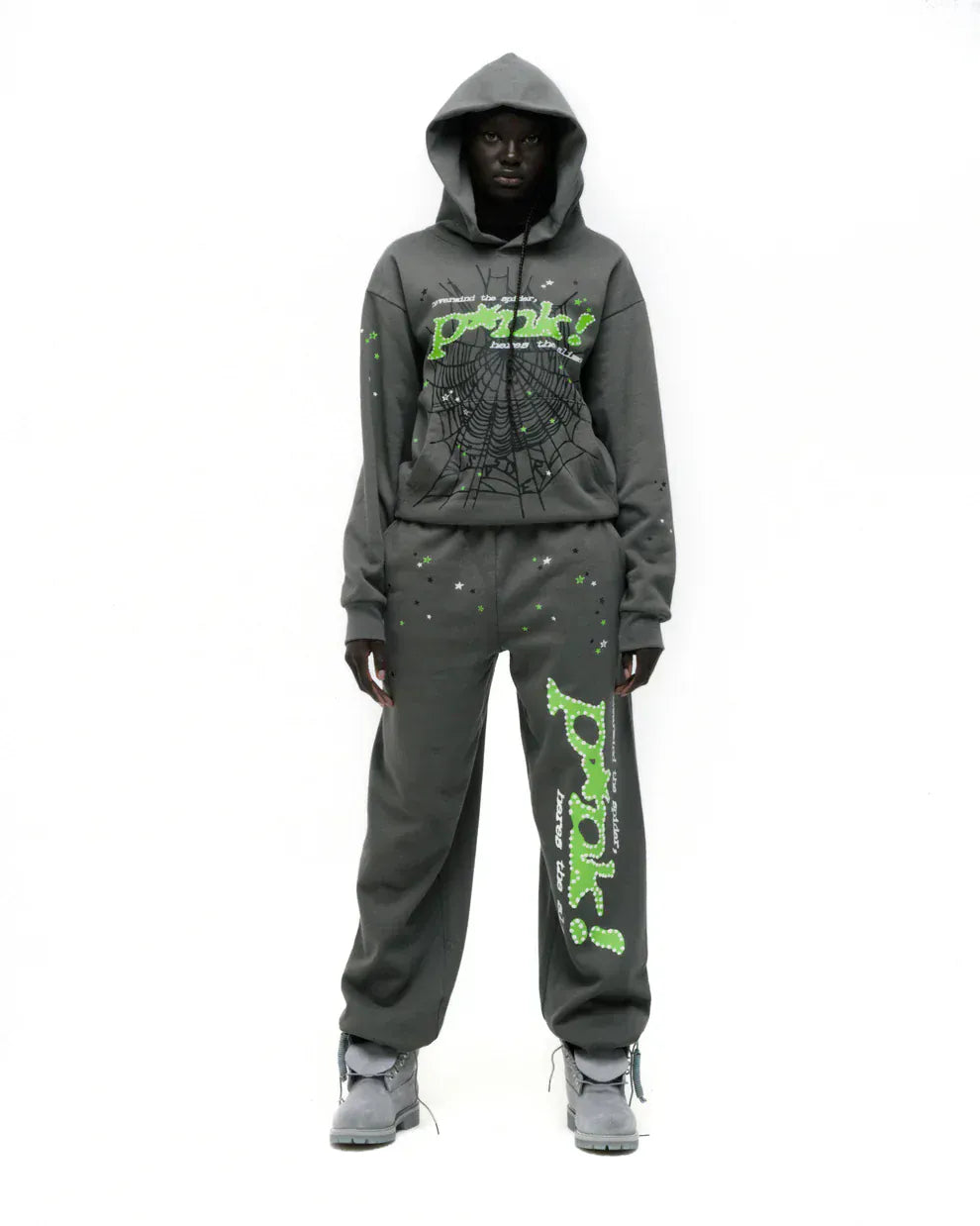 Wearing the Sp5der “P!nk! Hares the Slime” Hoodie in olive green, featuring bold spider-web graphics, and sweatpants with green graphic text, a person pairs gray sneakers for a striking streetwear look against a plain white background.