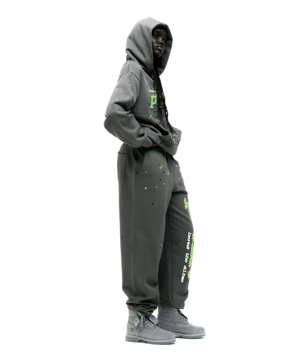Person wearing the Sp5der “P!nk! Hares the Slime” Olive Green 440GSM Heavyweight Spider-Web Pullover hoodie, paired with matching sweatpants and gray boots, strikes a bold streetwear pose against a white background.