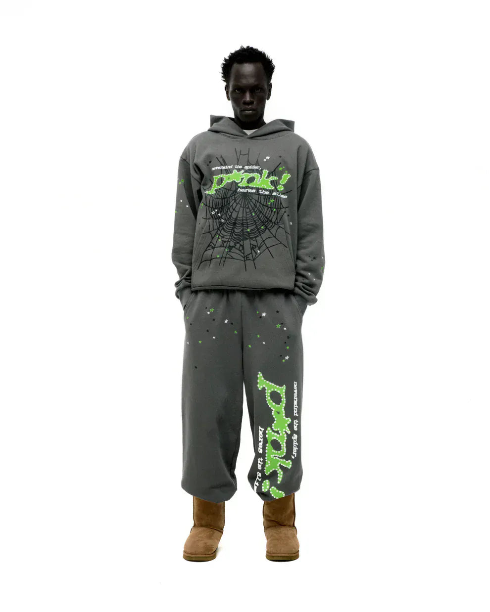 A person stands against a white background wearing an olive green Sp5der “P!nk! Hares the Slime” 440GSM heavyweight spider-web pullover hoodie, paired with matching pants and tan boots for a bold streetwear look.