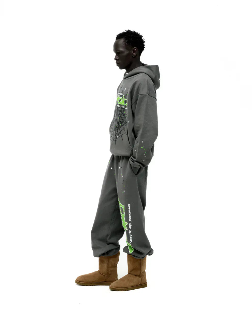 A person stands sideways in a Sp5der “P!nk! Hares the Slime” Olive Green 440GSM Heavyweight Spider-Web Pullover Hoodie with matching gray sweatpants with green graphics and tan boots, against a white background.