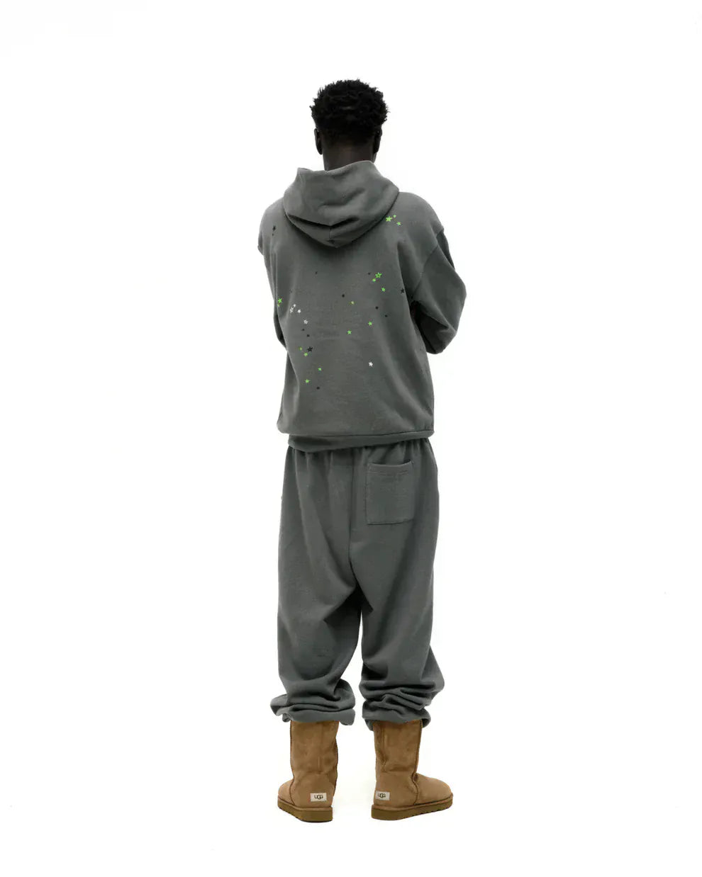 A person stands facing away, wearing an olive green Sp5der “P!nk! Hares the Slime” 440GSM heavyweight spider-web pullover hoodie with matching paint-splattered sweatpants and tan boots against a plain white background.