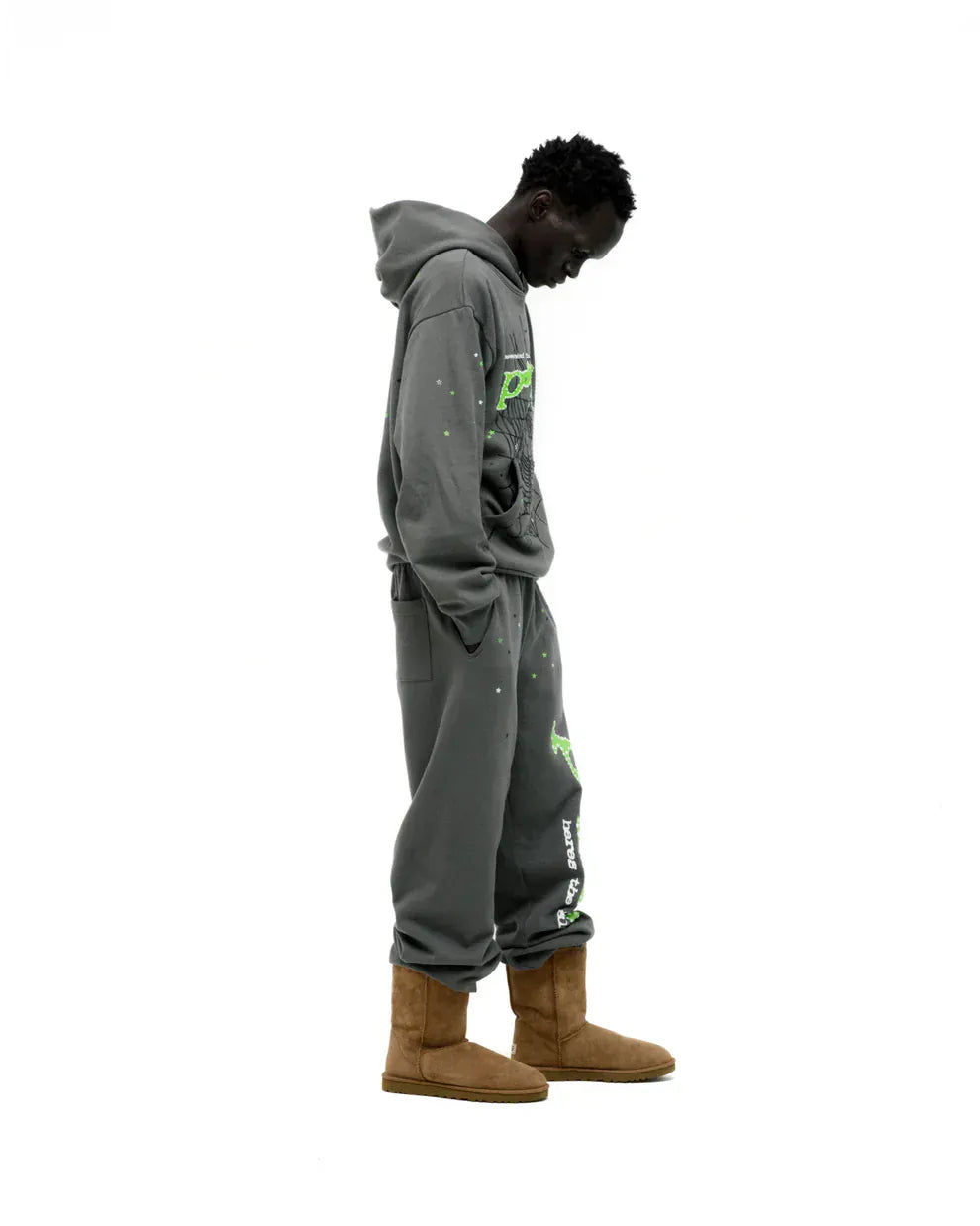A person faces left in the Sp5der “P!nk! Hares the Slime” Olive Green 440GSM Heavyweight Spider-Web Pullover, paired with sweatpants and tan boots, exuding streetwear vibes as they look down against a white background.