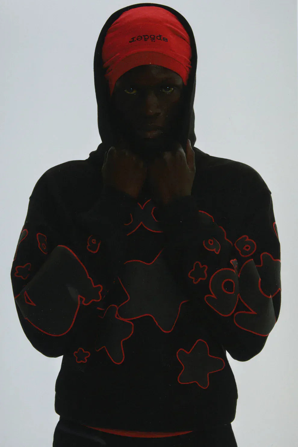 A person in a red head covering and the Sp5der 440GSM Heavyweight Star Hoodie – Black & Grey Oversized Streetwear Pullover stands facing forward, holding the hoodie collar and displaying bold streetwear style in heavyweight fleece.
