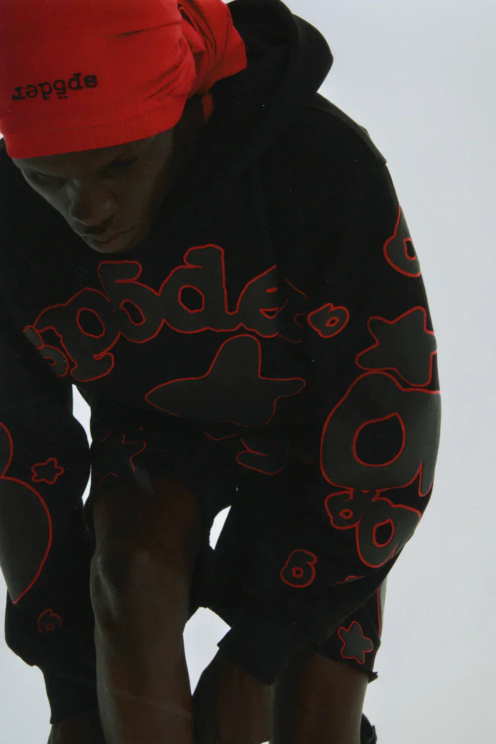 Wearing the Sp5der 440GSM Heavyweight Star Hoodie – Black & Grey Oversized Streetwear Pullover and matching shorts with red outlined graphics, a person in a red headscarf looks downward, embodying bold streetwear style against a plain backdrop.