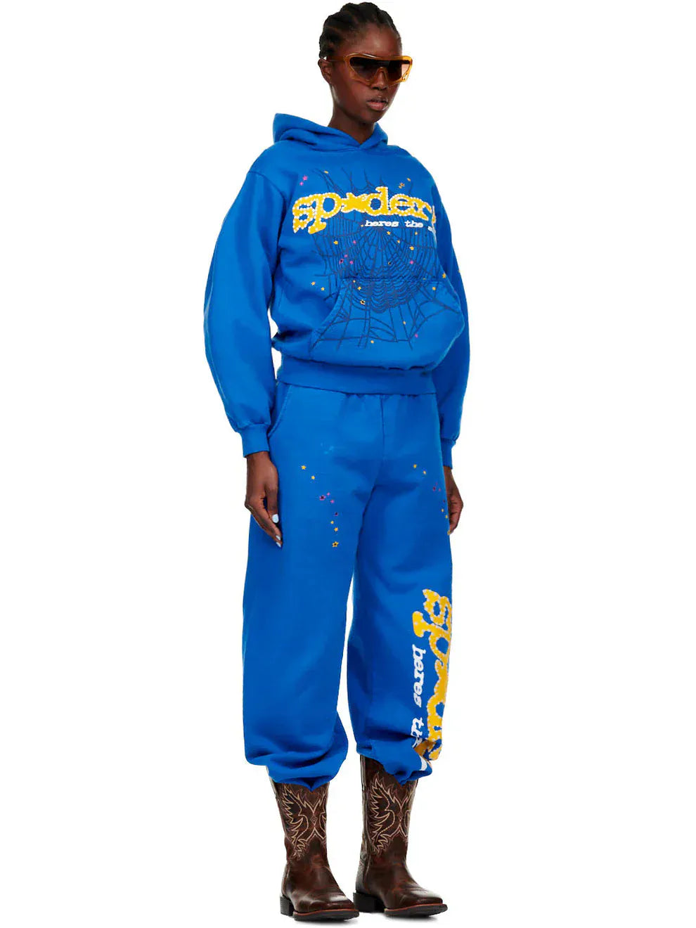 Person wearing the Sp5der “Hares the Slime” Hoodie (440GSM) in royal blue with a fuzzy spider-web graphic, matching sweatpants, brown cowboy boots, and sunglasses, standing against a white background.
