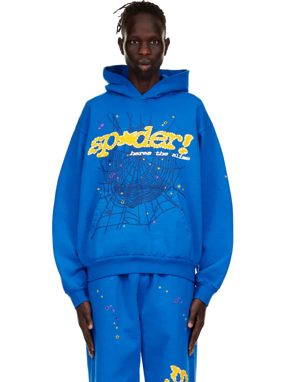 Someone rocks the Sp5der “Hares the Slime” Hoodie (440GSM) in royal blue, with a fuzzy logo and spider-web graphic—heavyweight streetwear that stands out.