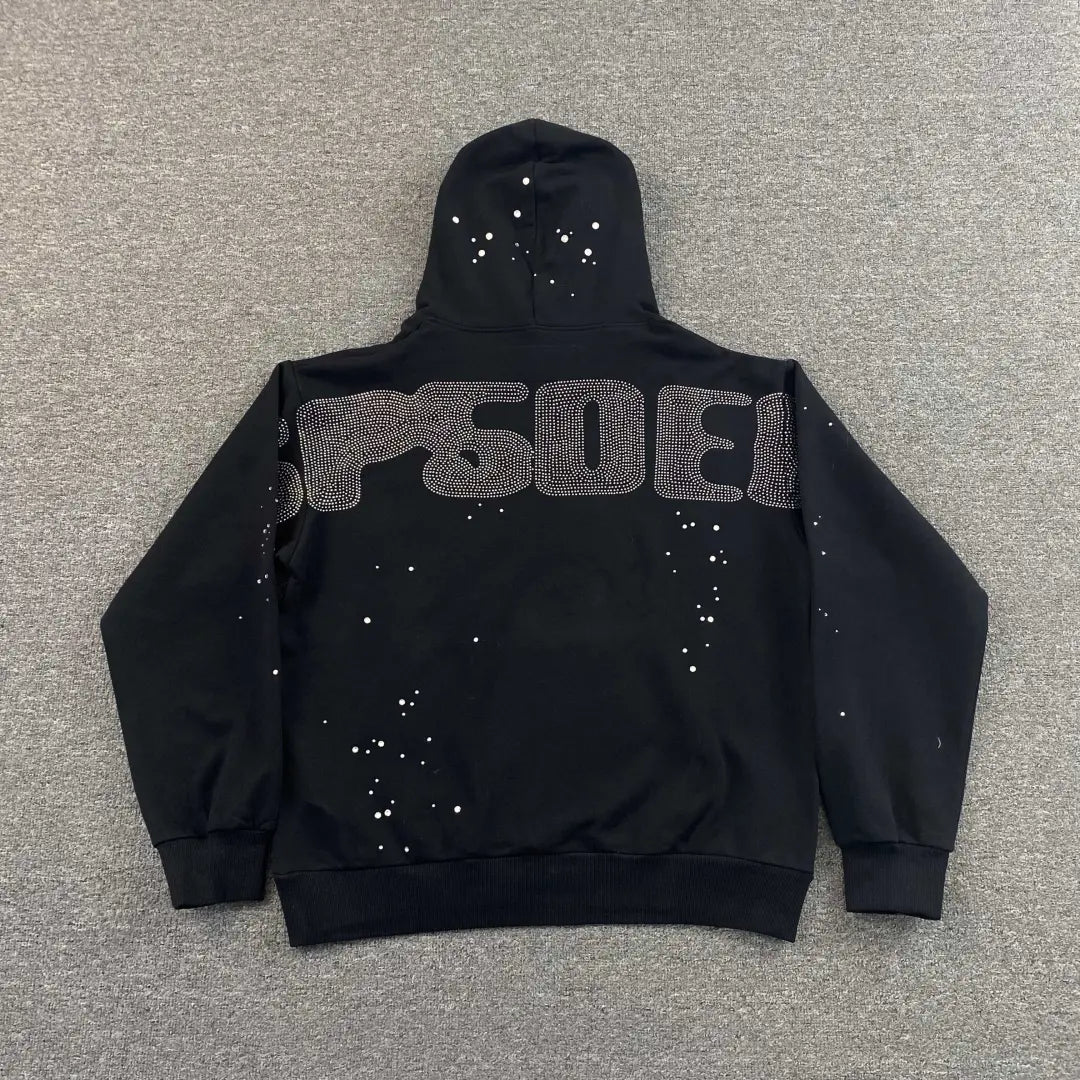 A black Sp5der Black Rhinestone Spiderweb Crystal Hoodie lies flat on gray, its back adorned with SPACE in large dotted letters and scattered white dots like stars, capturing the bold streetwear style.