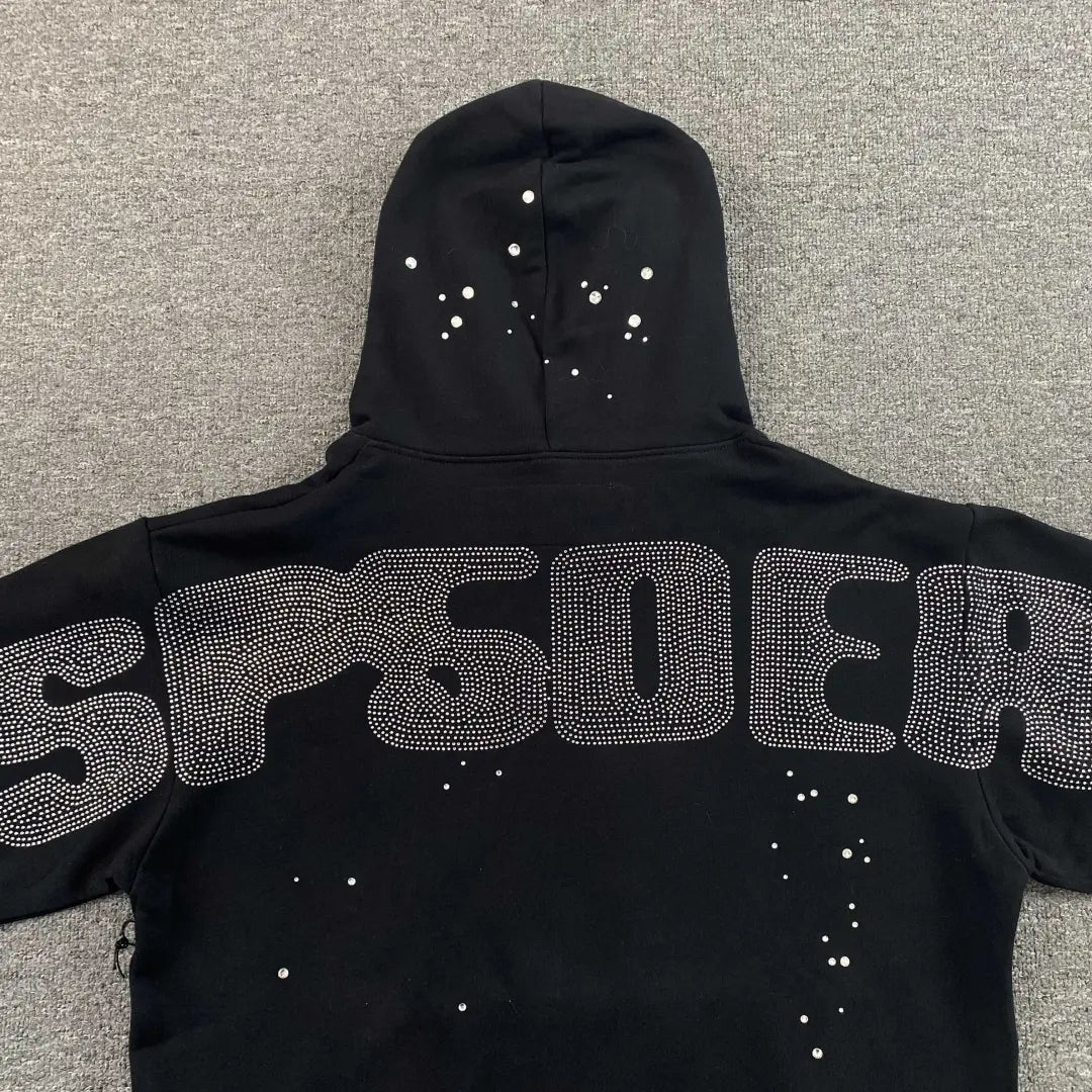 The Sp5der Black Rhinestone Spiderweb Crystal Hoodie features SP5DER in large silver-studded letters on the back and white spots on black fabric, laid flat on a gray surface―a bold streetwear essential.