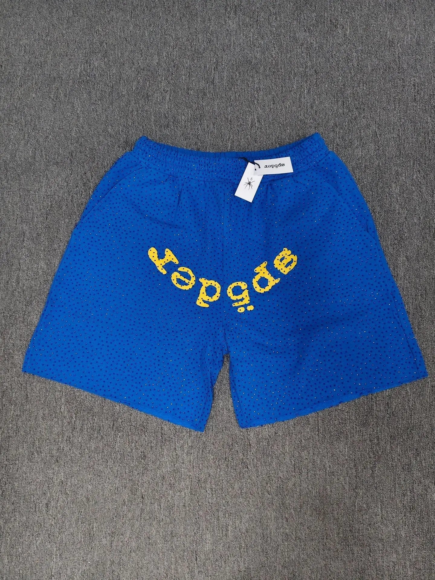 Blue Sp5der shorts with yellow front logo graphic and all-over dotted texture laid flat