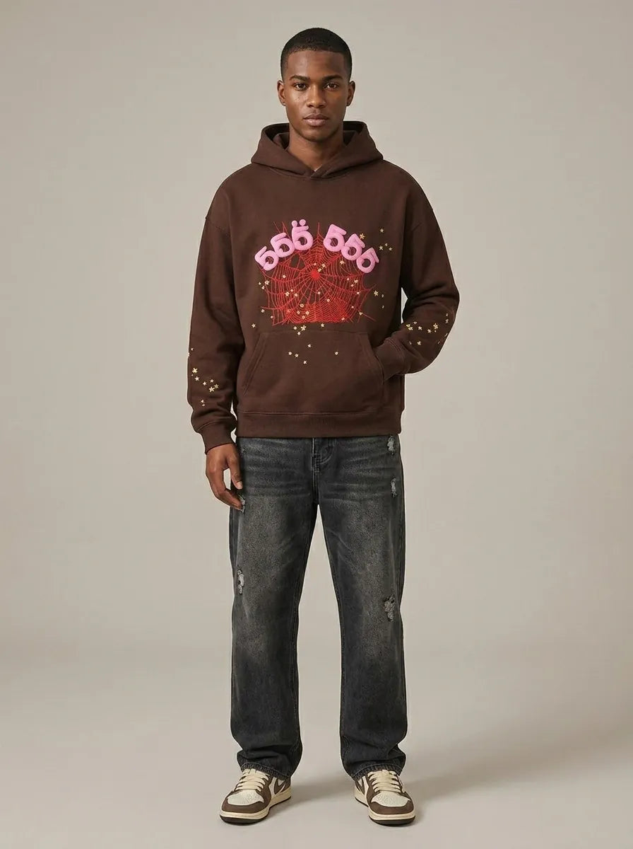 A man stands facing forward in the 555 Brown Spider Hoodie – Sp5der 555 Web Brown / Pink 440GSM Heavyweight Oversized Pullover, styled with distressed jeans and white sneakers, against a plain background.