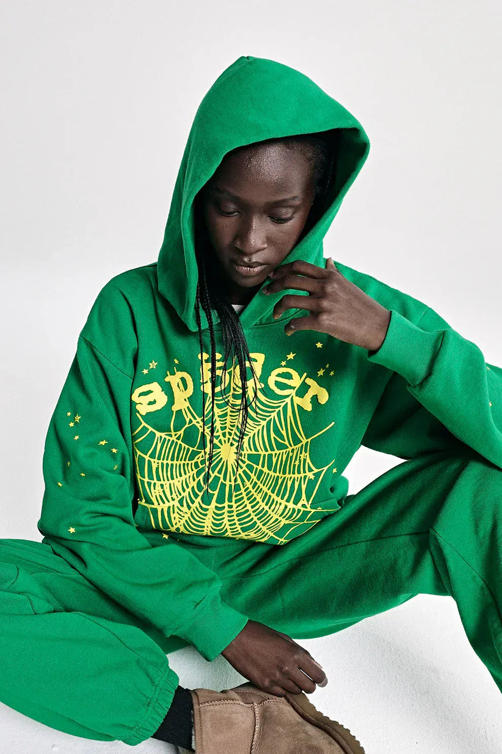 Wearing the Sp5der Web Hoodie – Emerald Green & Yellow 440GSM Heavyweight Oversized Streetwear Pullover, a person sits on the floor, hand on their hood, showcasing a cool streetwear vibe with matching pants and bold spider web designs.