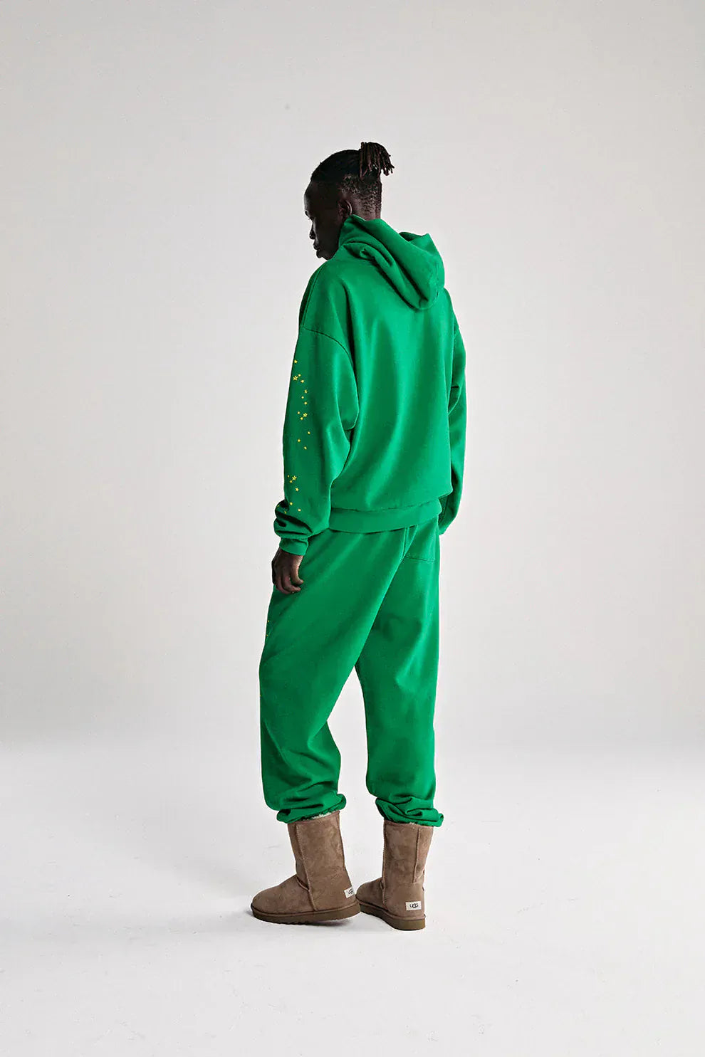 A person faces away in streetwear, wearing the Sp5der Web Hoodie – Emerald Green & Yellow 440GSM heavyweight oversized pullover and matching sweatpants with tan boots, set against a plain light background.