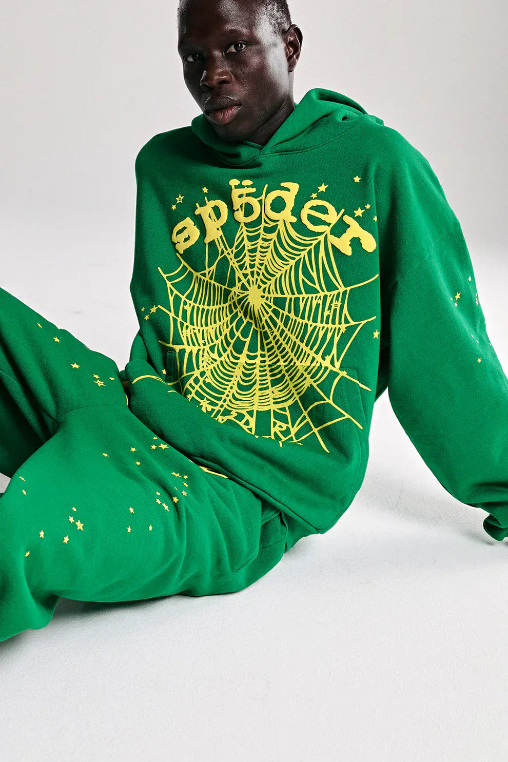 A person sits on a white surface wearing the Sp5der Web Hoodie – Emerald Green & Yellow 440GSM Heavyweight Oversized Streetwear Pullover with matching pants, featuring bold yellow web details—a standout streetwear look.