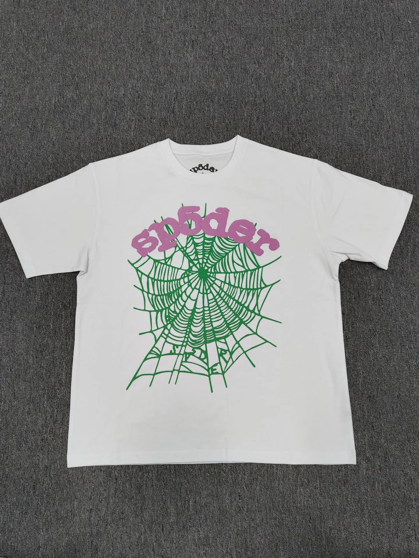 The Sp5der White Spider Web Graphic Cotton T-Shirt features a striking green spider web and bold pink "spider" text on the front, displayed flat on a dark gray surface to highlight its unique streetwear style.