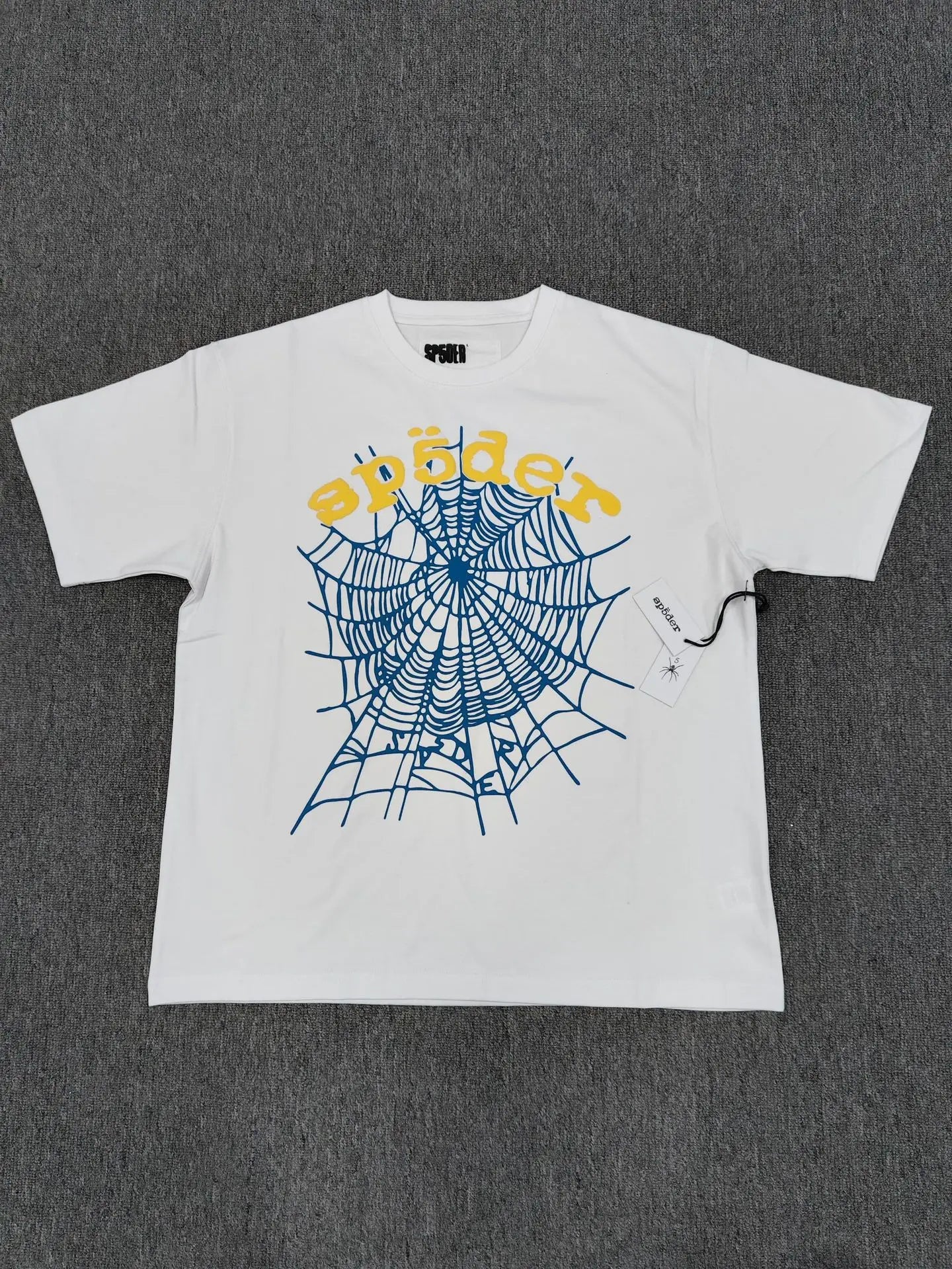 The Sp5der White Spider Web Graphic Cotton T-Shirt is laid flat on a gray surface, showcasing a bold blue spiderweb and yellow "sp5der" graphic across the front. A tag is attached to the right sleeve.