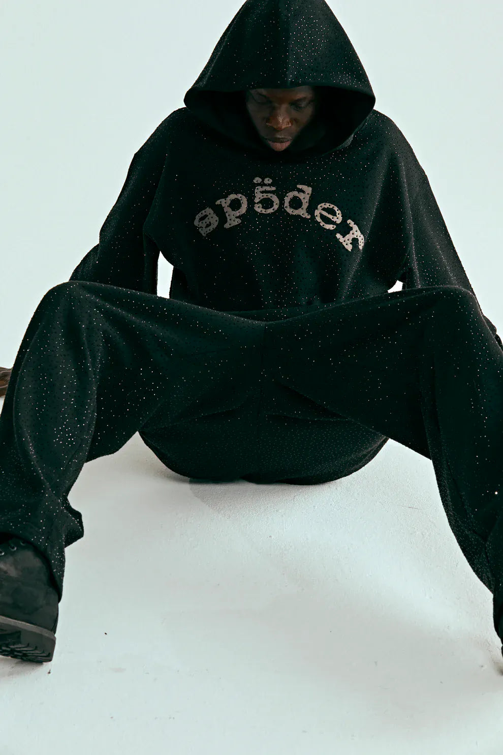 A person wearing the VVS Spider Hoodie – Black Sp5der Rhinestone Pullover with diamond graphic detail and white speckles on the front, sitting on the floor with legs spread and head slightly lowered.