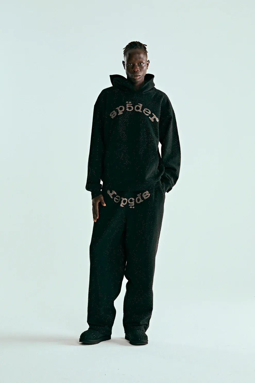 A person stands against a plain light background, wearing the VVS Spider Hoodie – Black Sp5der Rhinestone Pullover with Diamond Graphic Detail and matching black sweatpants featuring mirrored text on both pieces.