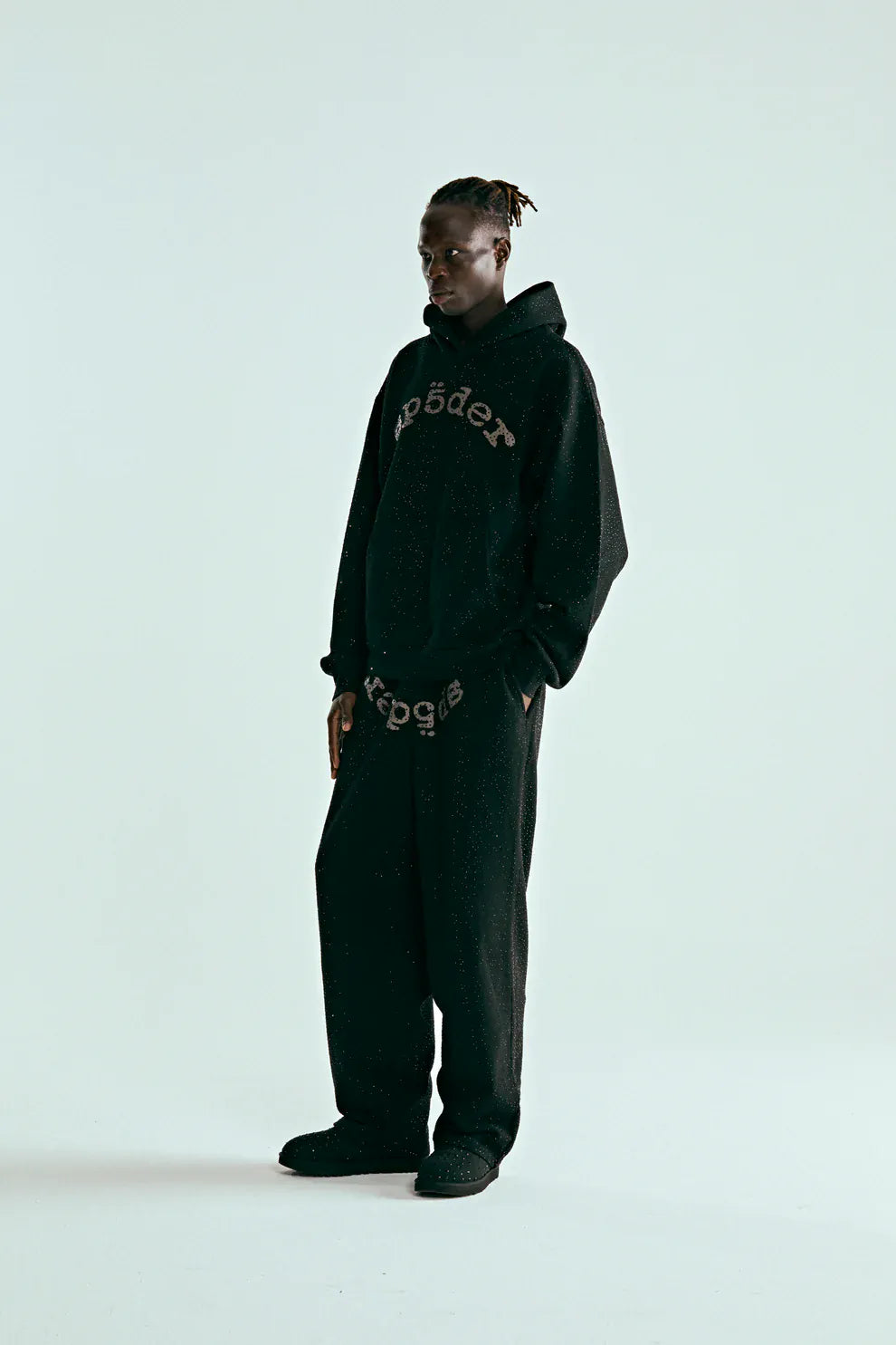 A person stands against a plain light background wearing the VVS Spider Hoodie – a black Sp5der rhinestone pullover with diamond graphic detail – paired with matching black pants featuring lettering and black shoes.