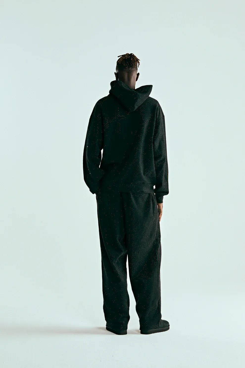 A person stands facing away, wearing the VVS Spider Hoodie – a black Sp5der rhinestone pullover with diamond graphic detail, paired with loose black pants against a plain light background.