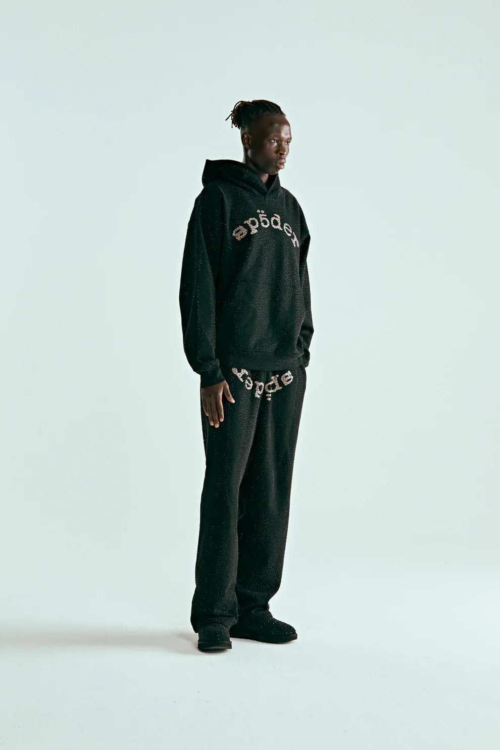 A person stands against a plain background wearing the VVS Spider Hoodie – a black Sp5der rhinestone pullover with diamond graphic detail and matching black sweatpants featuring patterned text across the front.