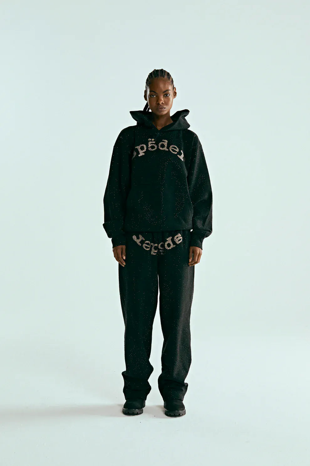 A person faces forward wearing the VVS Spider Hoodie – a black Sp5der rhinestone pullover with diamond graphic detail, paired with matching sweatpants, set against a plain light background.