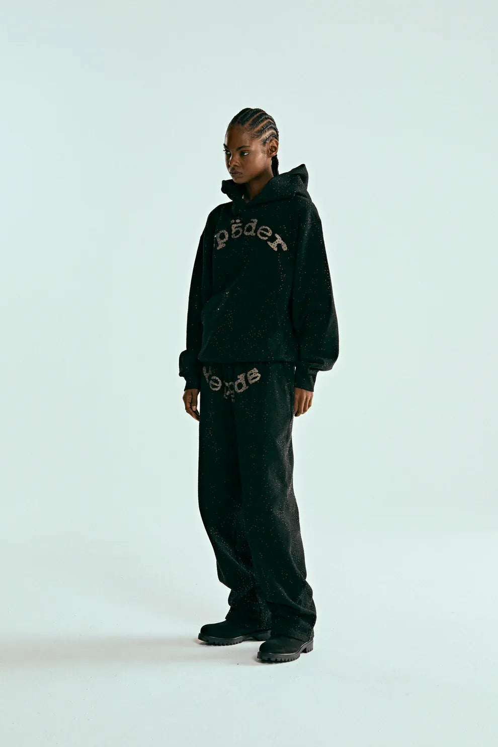 A person wears the VVS Spider Hoodie – Black Sp5der Rhinestone Pullover with diamond graphic detail, matching sweatpants featuring “SP5DER” print, and black shoes, standing against a plain light background.