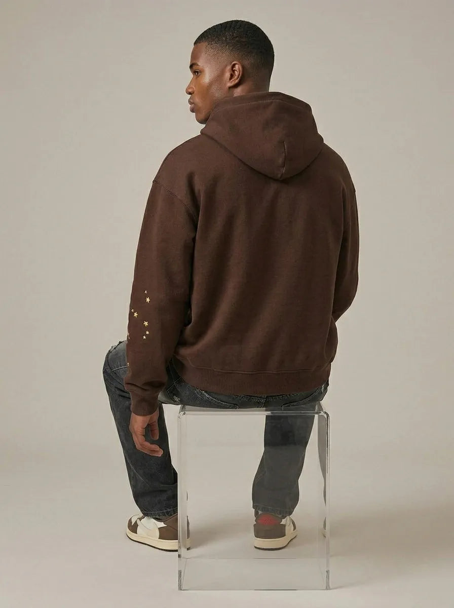 A man sits on a clear acrylic chair facing away, wearing the 555 Brown Spider Hoodie – Sp5der 555 Web Brown/Pink 440GSM Heavyweight Oversized Pullover, paired with dark jeans and white sneakers with brown and red accents.