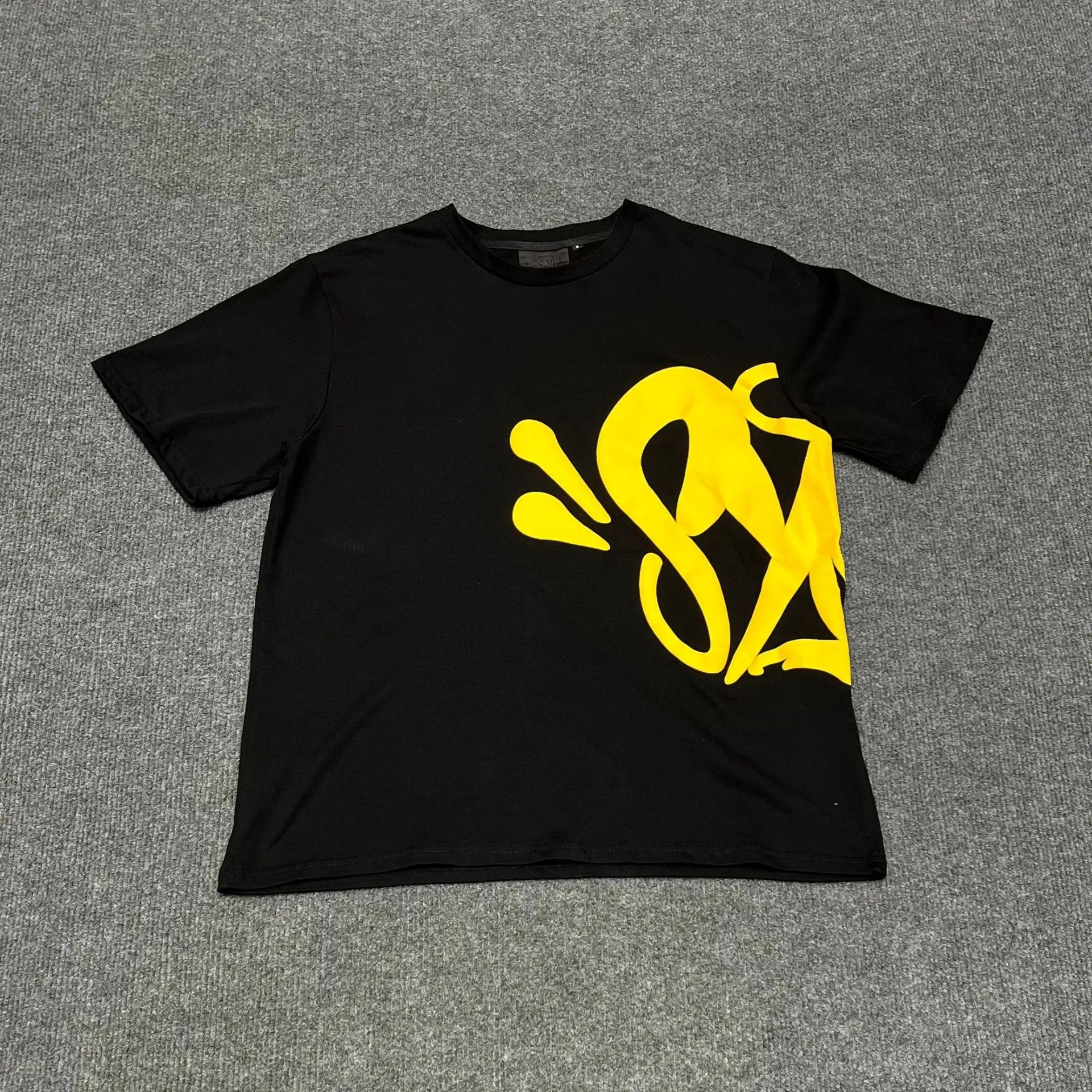 Black oversized T-shirt with large yellow graffiti graphic print on the front, laid flat on a grey surface